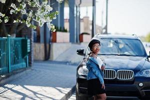 Stylish african american model in glasses hat, jeans jacket and black skirt posed outdoor against black business suv car. photo