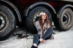 Brunette stylish casual girl in cap and sunglasses sitting against truck wheels. photo