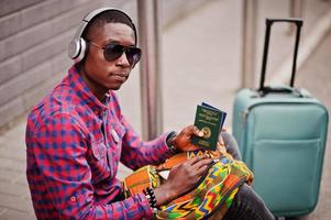 African american man in checkered shirt, sunglasses and earphones with suitcase and backpack holding Ghana passport. Black man traveler. photo