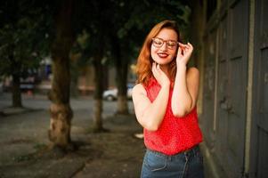 atractiva mujer pelirroja con anteojos, vestida con blusa roja y falda de jeans posando. foto