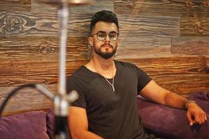 Stylish beard arabian man in glasses and black t-shirt smoking hookah indoor bar. Arab model having rest. photo