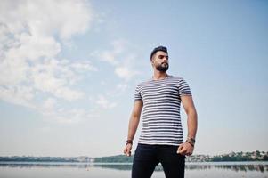 Handsome tall arabian beard man model at stripped shirt posed outdoor against lake and sky. Fashionable arab guy. photo