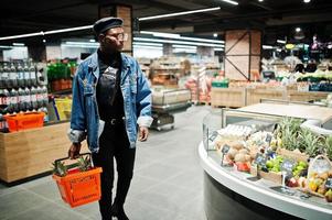 Elegante hombre afroamericano casual con chaqueta de jeans y boina negra sosteniendo una canasta con piñas en la sección orgánica de frutas del supermercado. foto