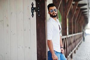 Stylish tall arabian man model in white shirt, jeans and sunglasses posed at street of city. Beard attractive arab guy. photo