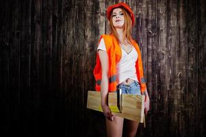 Engineer woman in orange protect helmet and building jacket against wooden background holding board and ruler. photo