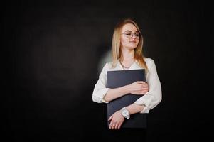 Studio portrait of blonde businesswoman in glasses, white blouse and black skirt holding laptop against dark background. Successful woman and stylish girl concept. photo