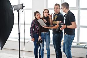 Photographer explaining about the shot to his team in the studio and looking on laptop. Talking to his assistants holding a camera during a photo shoot. Teamwork and brainstorm.