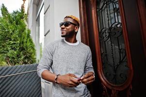 Elegante chico afroamericano con suéter gris y gafas de sol negras posadas en la calle con teléfono móvil. chico negro de moda. foto