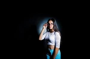 Portrait of an attractive young woman in white top and blue pants posing with her glasses in the dark. photo