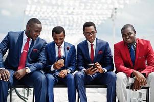 grupo de cuatro hombres afroamericanos felices y exitosos en traje. hombres de negocios negros ricos sentados en sillas mirando teléfonos móviles. foto