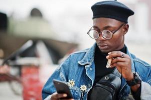 African american man in jeans jacket, beret and eyeglasses, smoking cigar and posed against old retro vehicle and looking at phone. photo