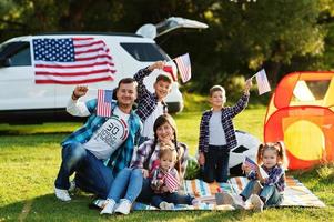 gran familia americana pasando tiempo juntos. con banderas de estados unidos contra gran coche suv al aire libre. vacaciones de américa. cuatro niños foto