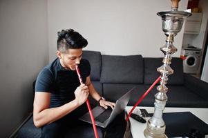 Handsome and fashionable indian man in black sitting at room, smoking hookah and working on laptop. photo