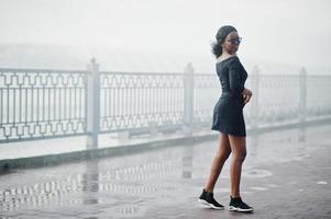 African american girl in sunglasses, black clothes and shirt posed outdoor. Fashionable black woman against rain of fountains. photo