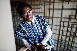 African businessman in traditional clothes and glasses with mobile phone at hand posed at modern building indoor. photo