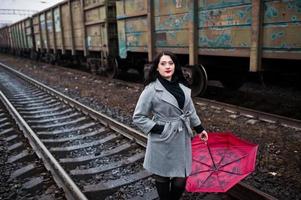chica morena con abrigo gris con paraguas rojo en la estación de tren. foto