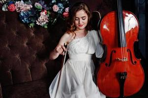 Pretty young gilrl musician in white dress with double bass sitting on brown vintage sofa. photo