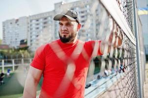 joven musculoso barbudo brutal vestido con camisa roja, pantalones cortos y gorra en el estadio. foto