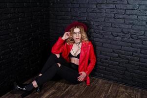 Studio portrait of blonde girl with red hat, glasses and leather jacket posed against brick wall. photo