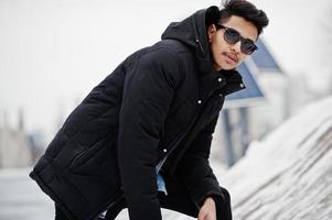 Casual young indian man in black jacket and sunglasses posed at winter day. photo