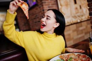 Funny brunette girl in yellow sweater eating pizza at restaurant. photo