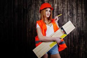Engineer woman in orange protect helmet and building jacket against wooden background holding board and ruler. photo