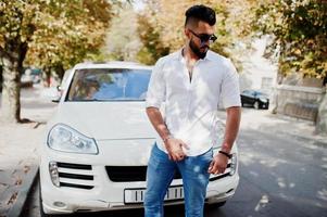 Elegante modelo de hombre árabe alto con camisa blanca, jeans y gafas de sol en la calle de la ciudad. chico árabe rico y atractivo con barba contra un auto todoterreno blanco. foto