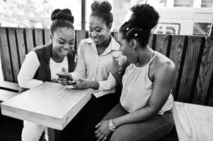 Three african american girls sitting on the table of caffe and looking on mobile phone. photo