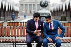 Two elegant indian fashionable mans model on suit posed at winter day sitting on bench and looking at phone. photo