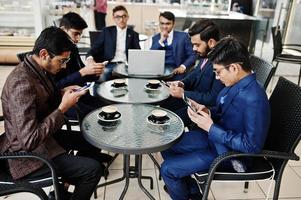 Group of six indian business man in suits sitting at office on cafe,  looking at laptop, texting on phones and drinking coffee. photo