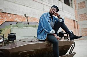 African american man in jeans jacket, beret and eyeglasses, smoking cigar and posed against btr military armored vehicle. photo