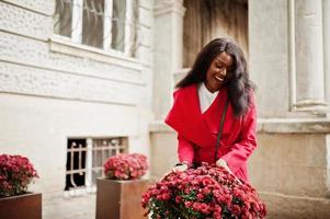 elegante mujer afroamericana con abrigo rojo posada contra flores al aire libre. foto