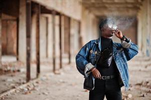 hombre afroamericano con chaqueta de jeans, boina y anteojos, fumando cigarro en una fábrica abandonada. foto