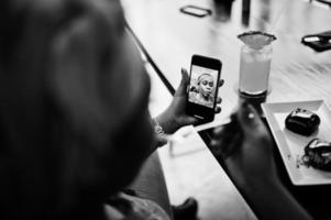 Stylish african woman in red shirt and hat posed indoor cafe, eat chocolate dessert cakes and making selfie at mobile phone. photo