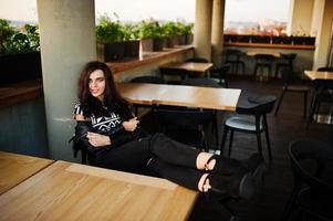 Young curly woman in leather jacket in a bar put her legs on table. photo