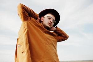 Stylish man in glasses, brown jacket and hat posed on green field. photo