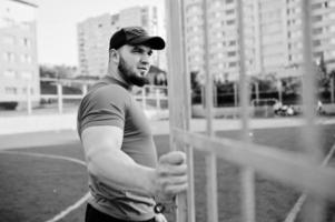 joven musculoso barbudo brutal vestido con camisa roja, pantalones cortos y gorra en el estadio. foto