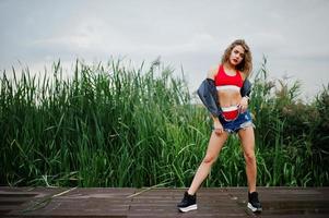 Sexy curly model girl in red top, jeans denim shorts, leather jacket and sneakers posed in the reed. photo