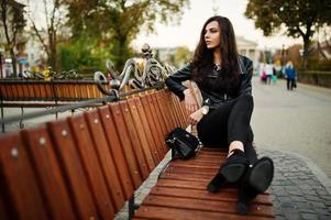 Young curly and sexy woman in leather jacket sitting on bench at street. photo