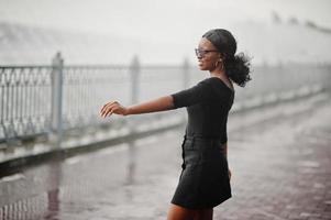 African american girl in sunglasses, black clothes and shirt posed outdoor. Fashionable black woman against rain of fountains. photo