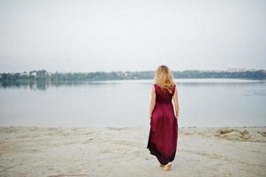 Back of blonde sensual barefoot woman in red marsala dress posing against lake. photo
