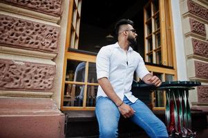 elegante modelo árabe alto con camisa blanca, jeans y gafas de sol sentado en la calle de la ciudad. barba chico árabe atractivo contra la ventana abierta del restaurante al aire libre. foto