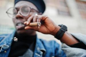 Close up portrait of african american man in jeans jacket, beret and eyeglasses, smoking cigar. photo