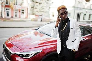 Chic handsome african american man in white suit against red luxury car with mobile phone at hand. photo