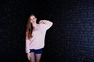 Studio shot of brunette girl at pink sweater with jeans shorts against black brick wall. photo