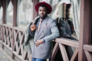 Stylish African American man model in gray coat, jacket tie and red hat posed against wooden cafe. photo