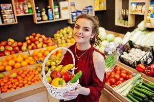 Girl in red holding different fruit and vegetables at basket on fruits store. photo