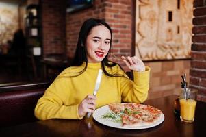 Funny brunette girl in yellow sweater eating pizza at restaurant. photo