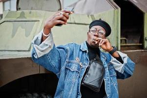 African american man in jeans jacket, beret and eyeglasses, smoking cigar and posed against btr military armored vehicle, making selfie on phone. photo