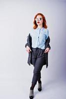 Portrait of a fantastic redheaded girl in blue shirt, grey overcoat posing with sunglasses in the studio. photo